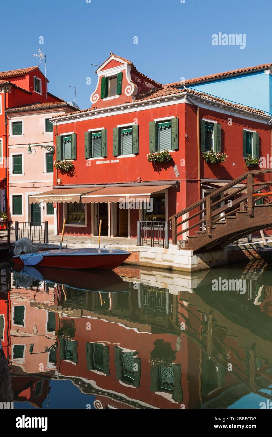 Brown wooden footbridge over canal with moored boat and pink and red stucco houses and a storefront, Burano Island, Venetian Lagoon, Venice, Veneto, Italy Stock Photo