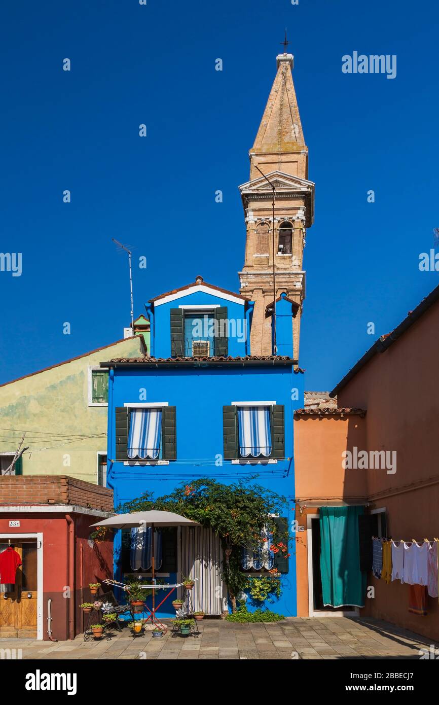 Blue stucco house facade decorated with striped curtains over entrance door and windows, leaning bell tower of San Martino church, Burano Island, Venetian Lagoon, Venice, Veneto, Italy Stock Photo