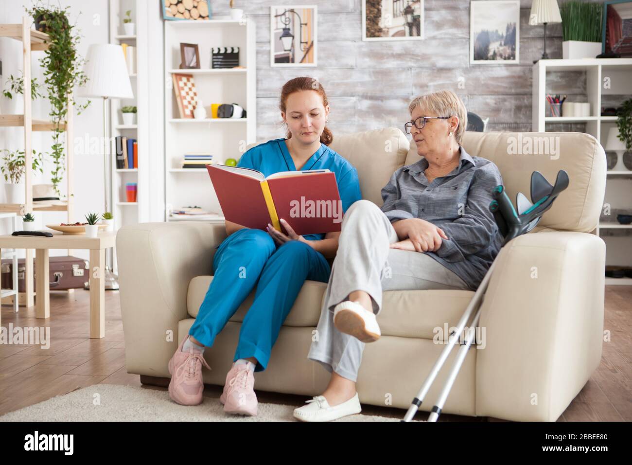 Young nurse sitting on couch in nursing home reading a book for old