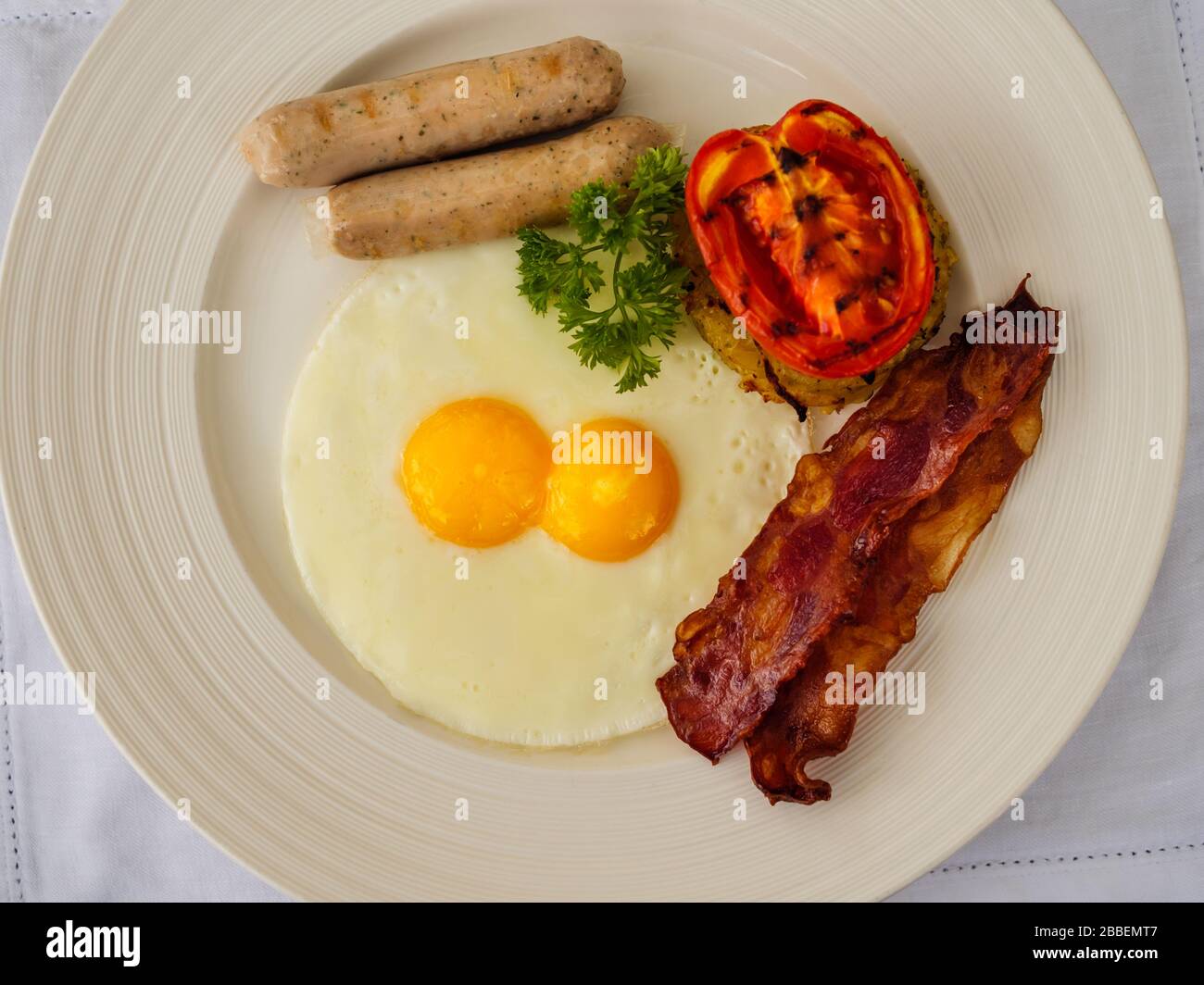 Flat lay view of a western fried breakfast – bacon and eggs, sausages and fried tomato on white plate and white tablecloth with copy space Stock Photo