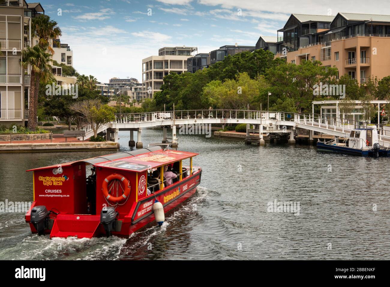 South Africa, Western Cape, Cape Town, Victoria and Alfred Waterfront, red City Sightseeing cruise boat passing through upmarket waterside housing on Stock Photo