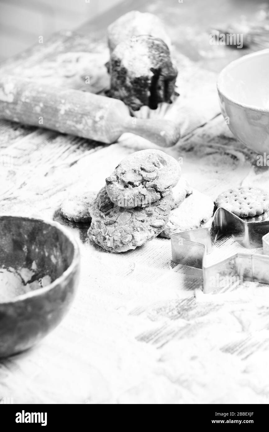 tasty delicious chip chocolate cookies sprinkled with powdered sugar near red turquoise bowls, cutter and rolling pin on wooden table with lot of flour, selective focus Stock Photo