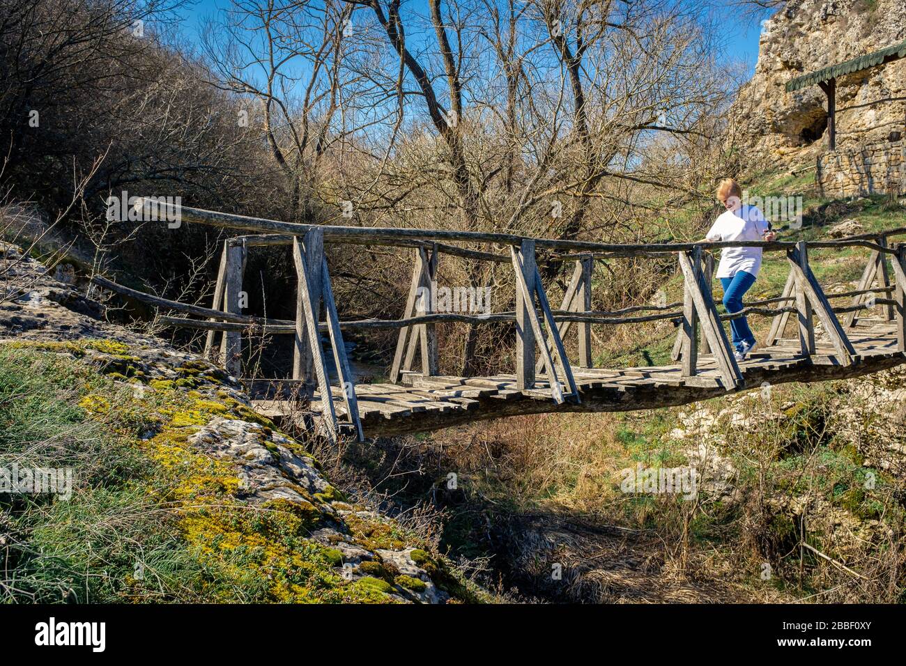 Caucasian women crossing over an old unreliable rickety man made wooden foot bridge over a river gorge. Emen gorge Bulgaria Stock Photo
