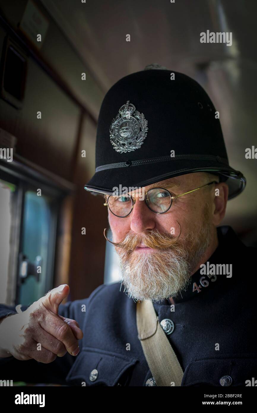 Isolated 1940s man with handlebar moustache as vintage UK policeman wearing helmet on board vintage steam train inside heritage railway carriage, Britain. Stock Photo