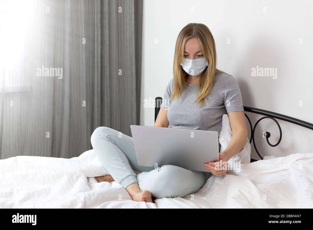young sick woman in a medical mask and a gray t-shirt and comfortable pants sitting on a white bed with a laptop . she works from home because of the Stock Photo