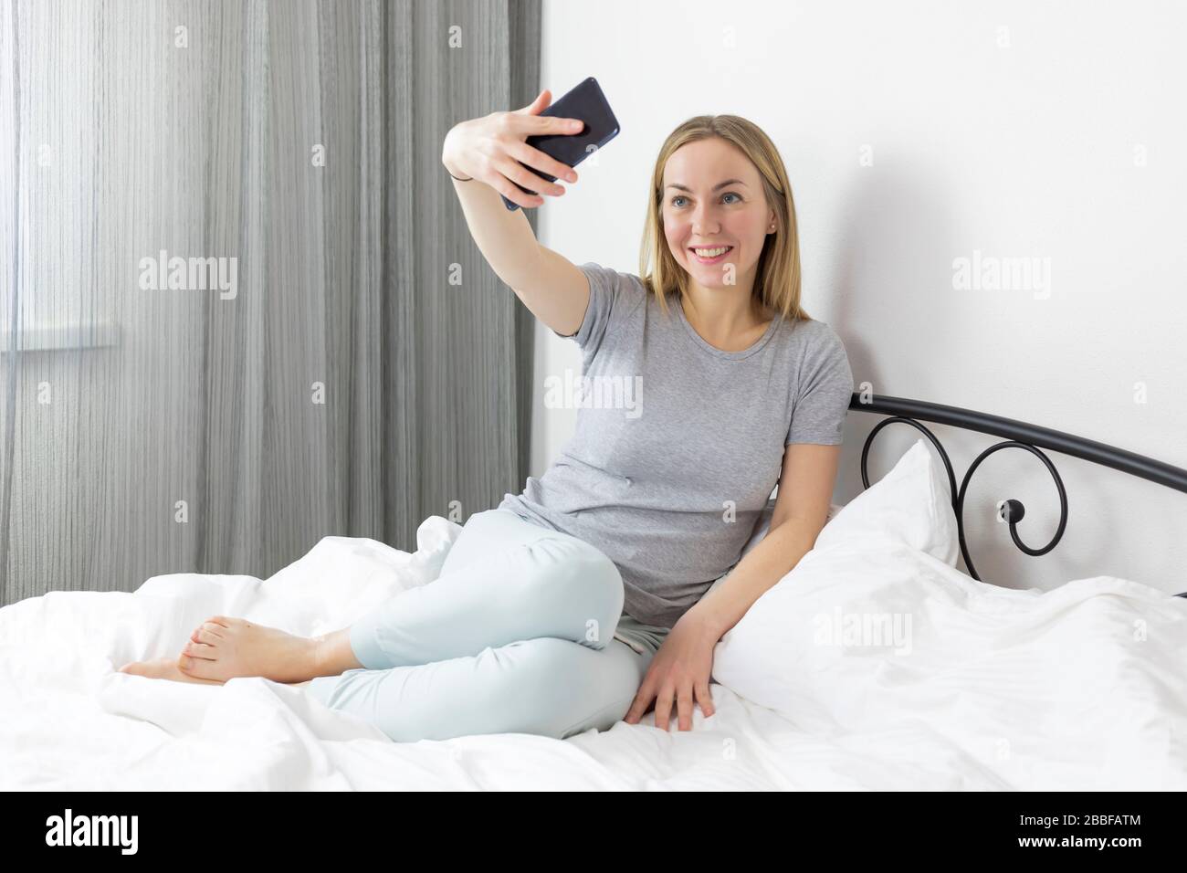 Young pretty european blonde woman working and studying from home, makes selfie and smiles, laughs . in bed with laptop and smartphone. she is in ligh Stock Photo