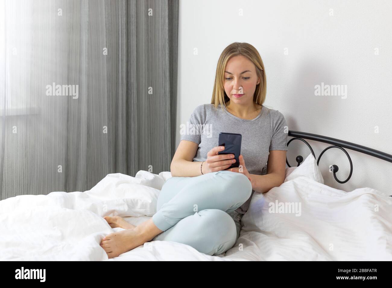 Young pretty european blonde woman working and studying from home . in bed with smartphone. she is in light, white, comfortable home clothes. Stock Photo