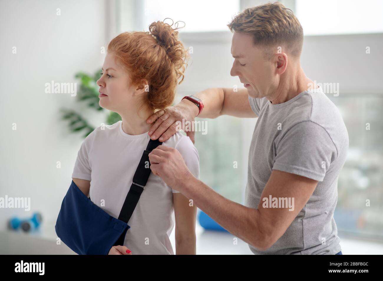 Male physiotherapist examining shoulder of female patient with arm sling Stock Photo