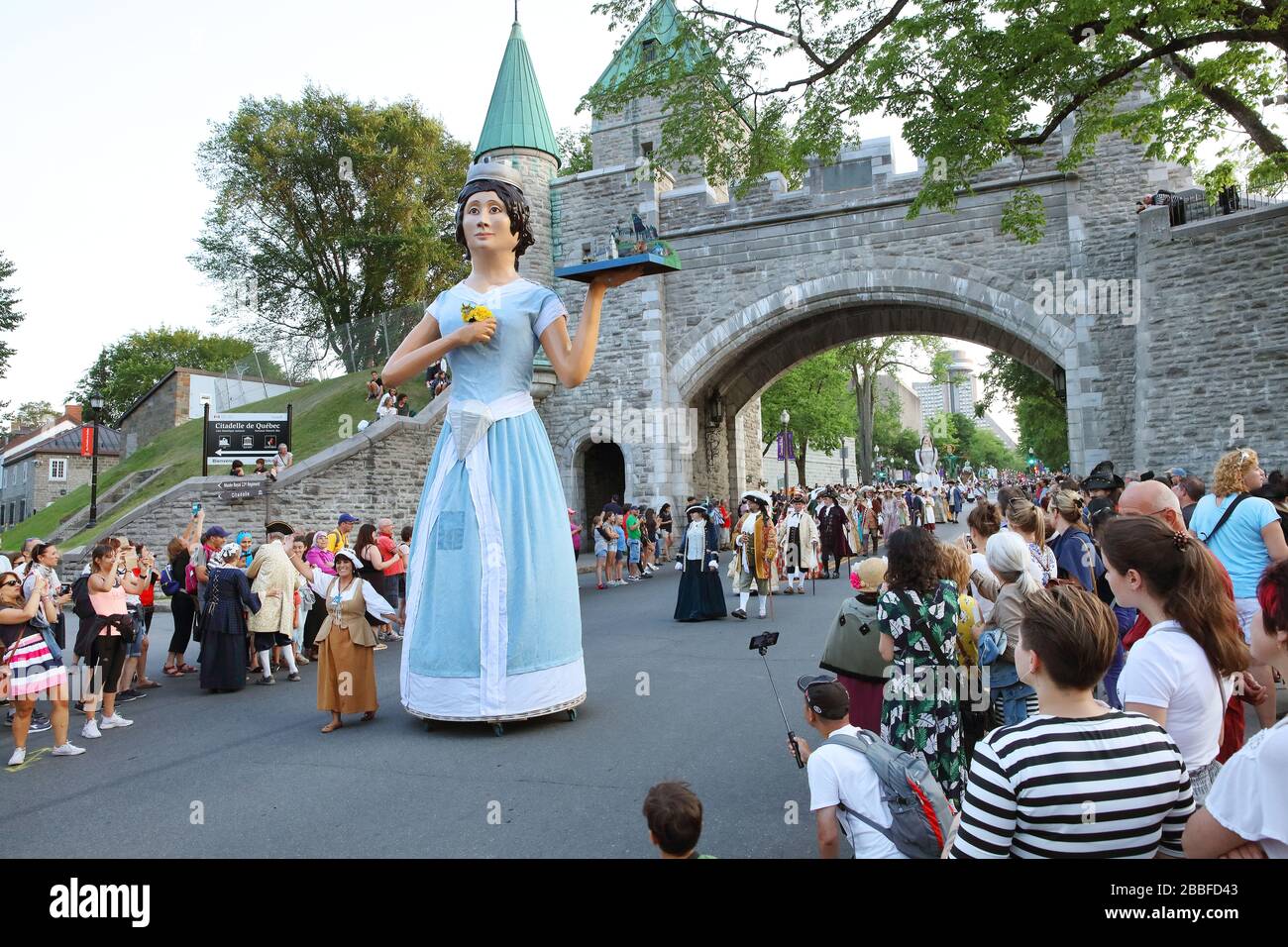 Parade with participants in period costumes and 'giants' symbolizing historical characters or locations when Quebec City was part of New France (late 1800s). The giant in the photo is of Dame Cap-Diamant (Lady Cape Diamond) and, in the background, is Porte Saint-Louis, Quebec City, Province of Quebec, Canada Stock Photo