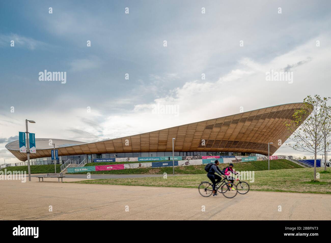 A couple of cyclists ride past the architecturally impressive cycling center and velodrome used during the 2012 summer olympics in London Stock Photo