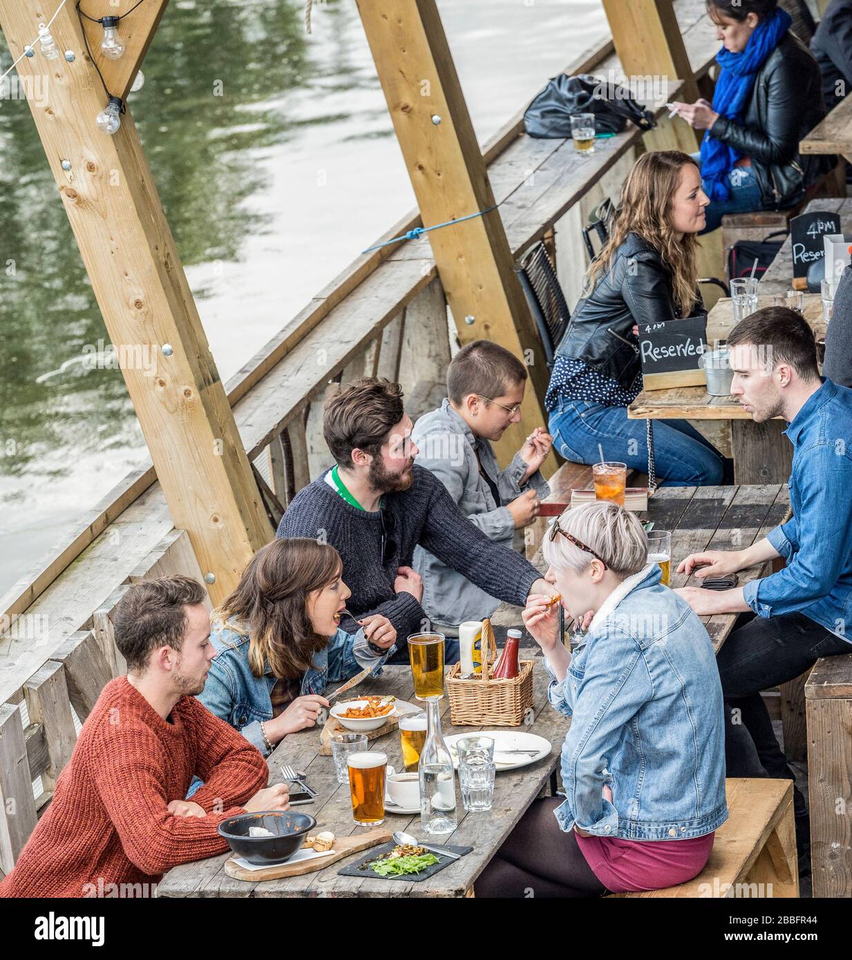 Groups of young hipsters socialise on a riverside bar and restaurant terrace on the bank of the river lea in Hackney Wick, East London, uk Stock Photo