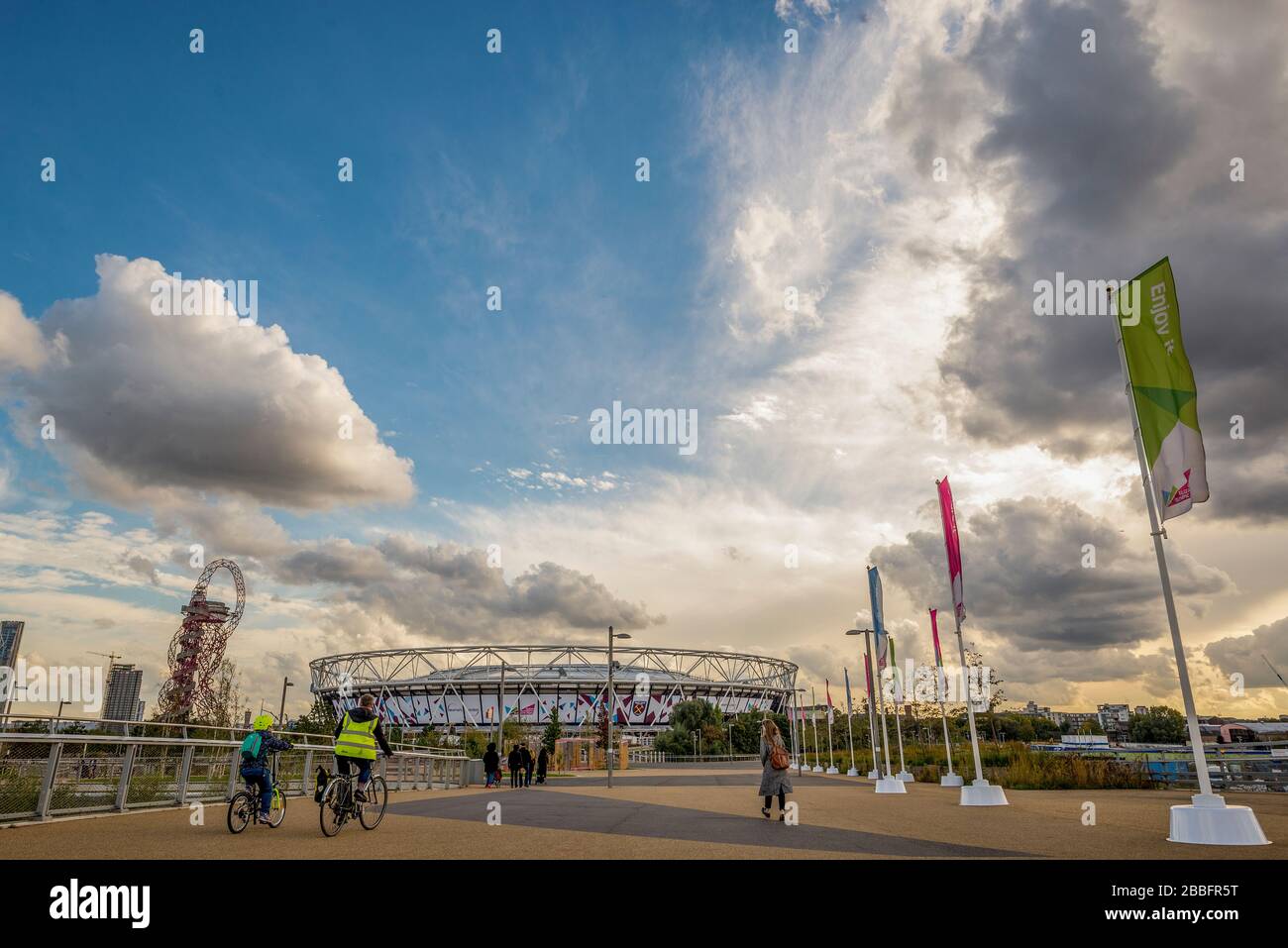 The sun sets over the 2012 Olympic stadium now home to football team West Ham United. People walk and cycle on the path that leads to the ground. Stock Photo