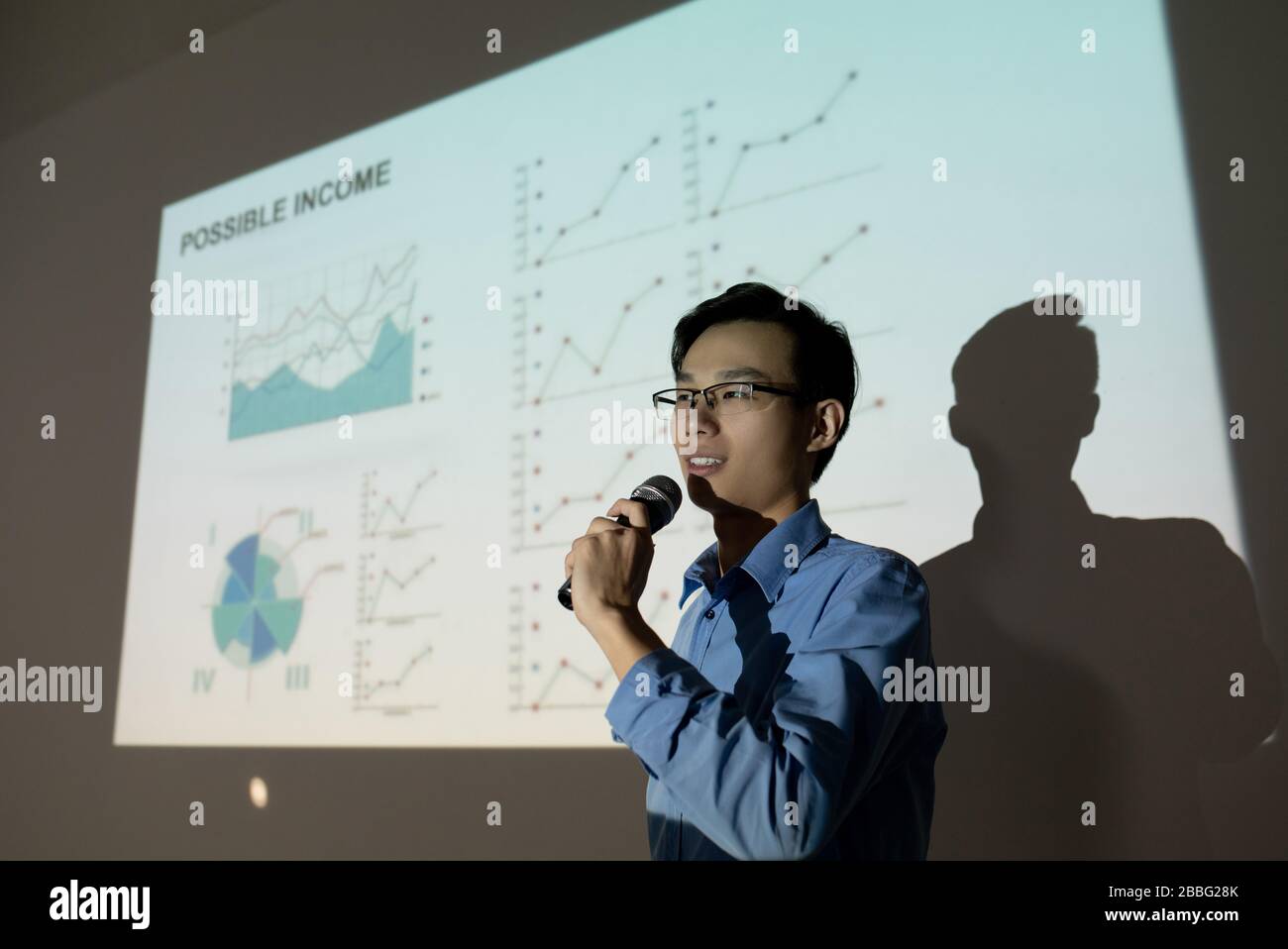 Confident young Asian man in eyeglasses standing at projection screen and speaking into microphone while presenting his business idea to investors Stock Photo