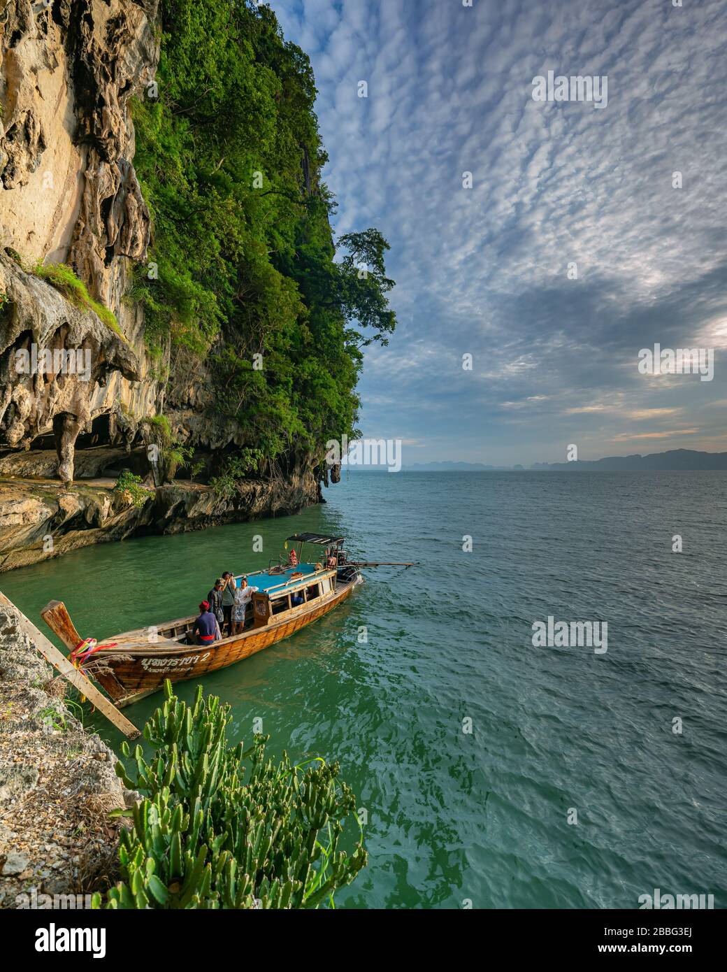 Thai long tail boat with tourists moored to a limestone cliff island with emerald green sea in Krabi district, Thailand. Stock Photo