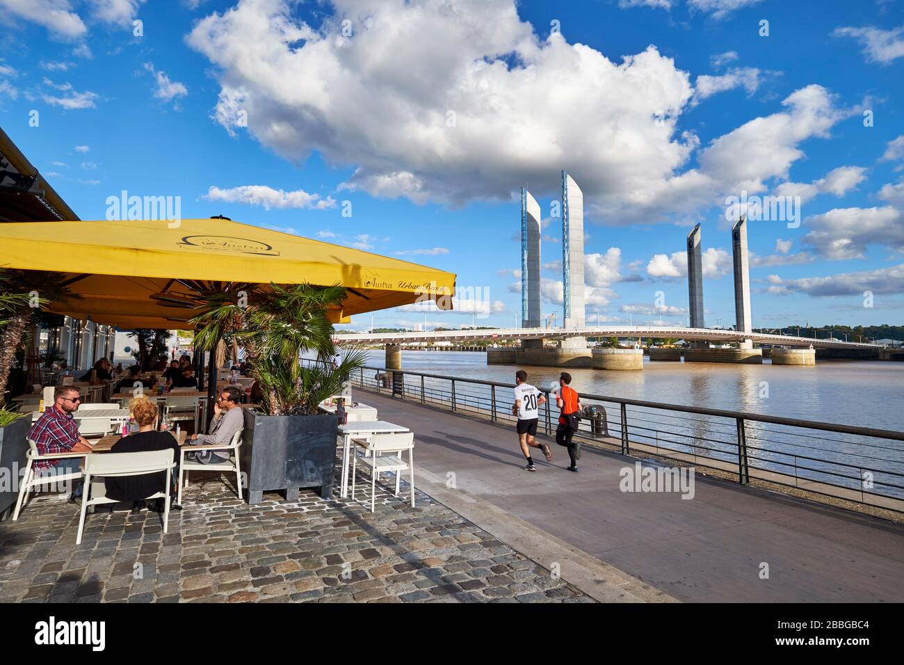 View of the Jacques Chaban-Delmas vertical-lift bridge on the Garonne river with café terrace and two male's running on the riverfront walkway Stock Photo