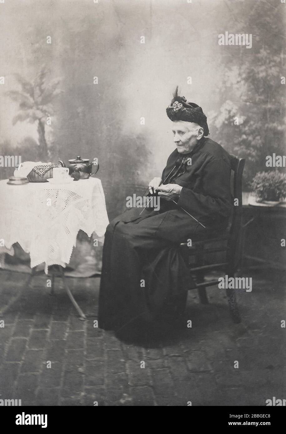 studio photo setup showing an old woman stitching, knitting, sewing in front of a typical studio background around 1900-1910 in the area of Antwerp, B Stock Photo