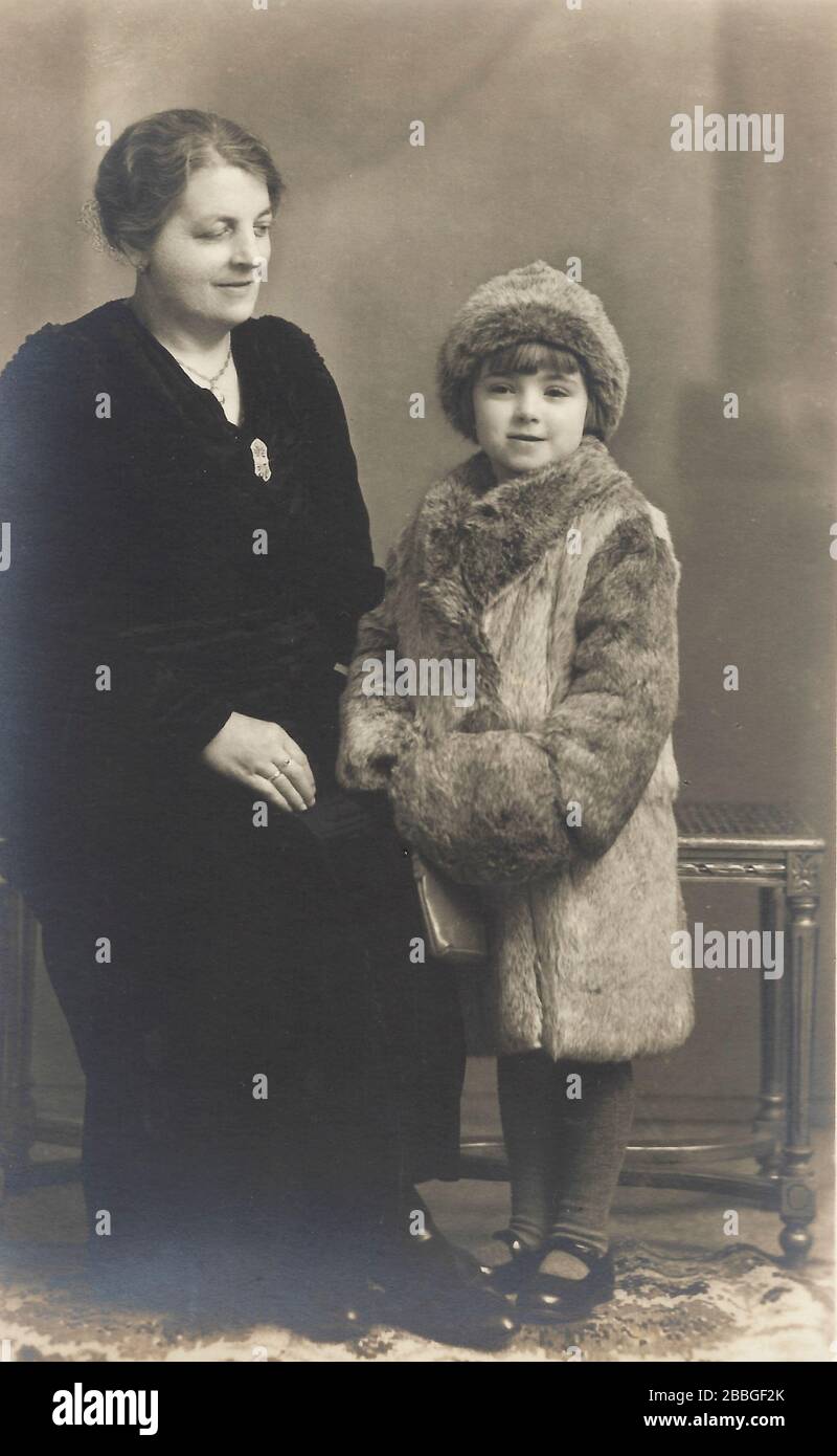 A vintage photo taken in studio around 1900-19100, showing a young 8-10 year old girl with her mother posing with an adult real fur coat outfit clothi Stock Photo