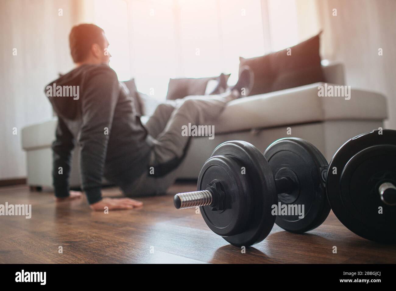 Young ordinary man go in for sport at home. Hardworking beginner sit on floor and hold legs on sofa. Freshmen trying to do abs exercising. Home alone Stock Photo