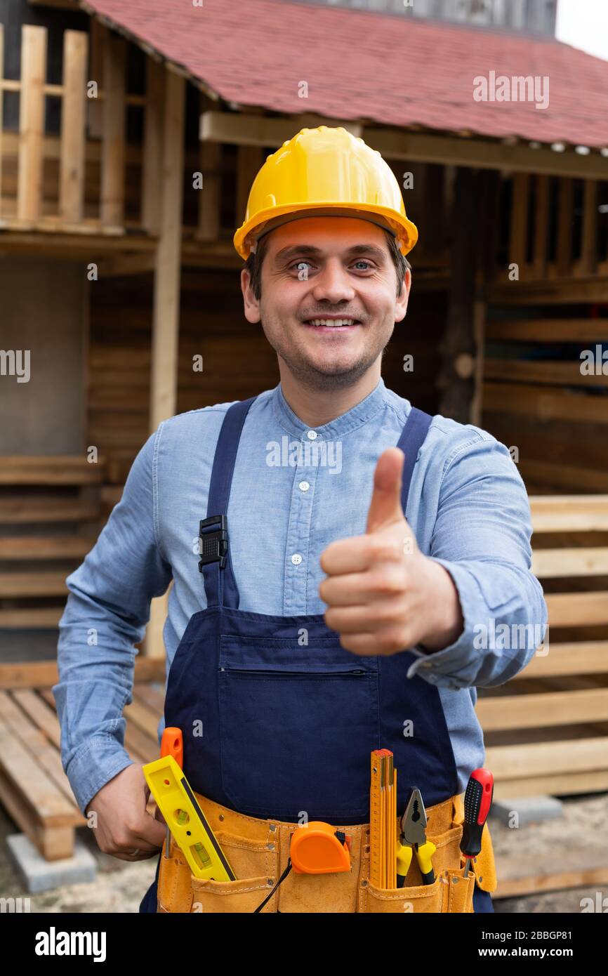 Young carpenter with yellow hard hat and work equipment showing thumbs