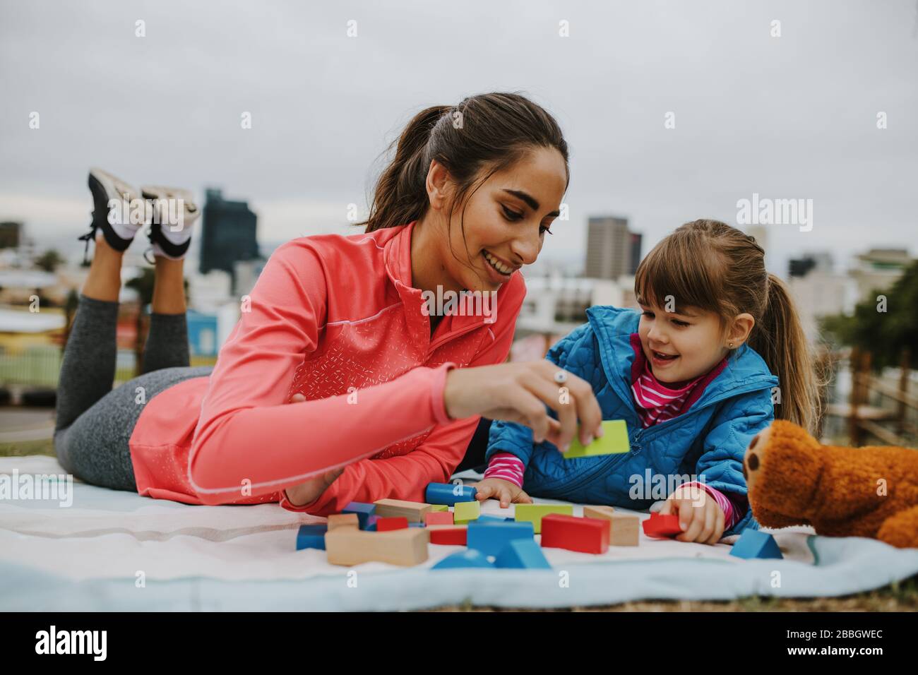 Woman playing building blocks with a small girl at the park. Girl child and her nanny lying on the blanket and playing wooden blocks at the park. Stock Photo