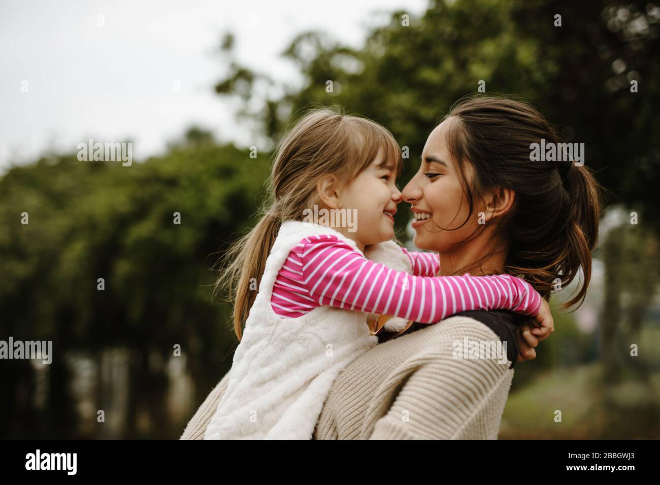 Side view of woman carrying a small girl and smiling outdoors. Babysitter with a girl at the park. Stock Photo