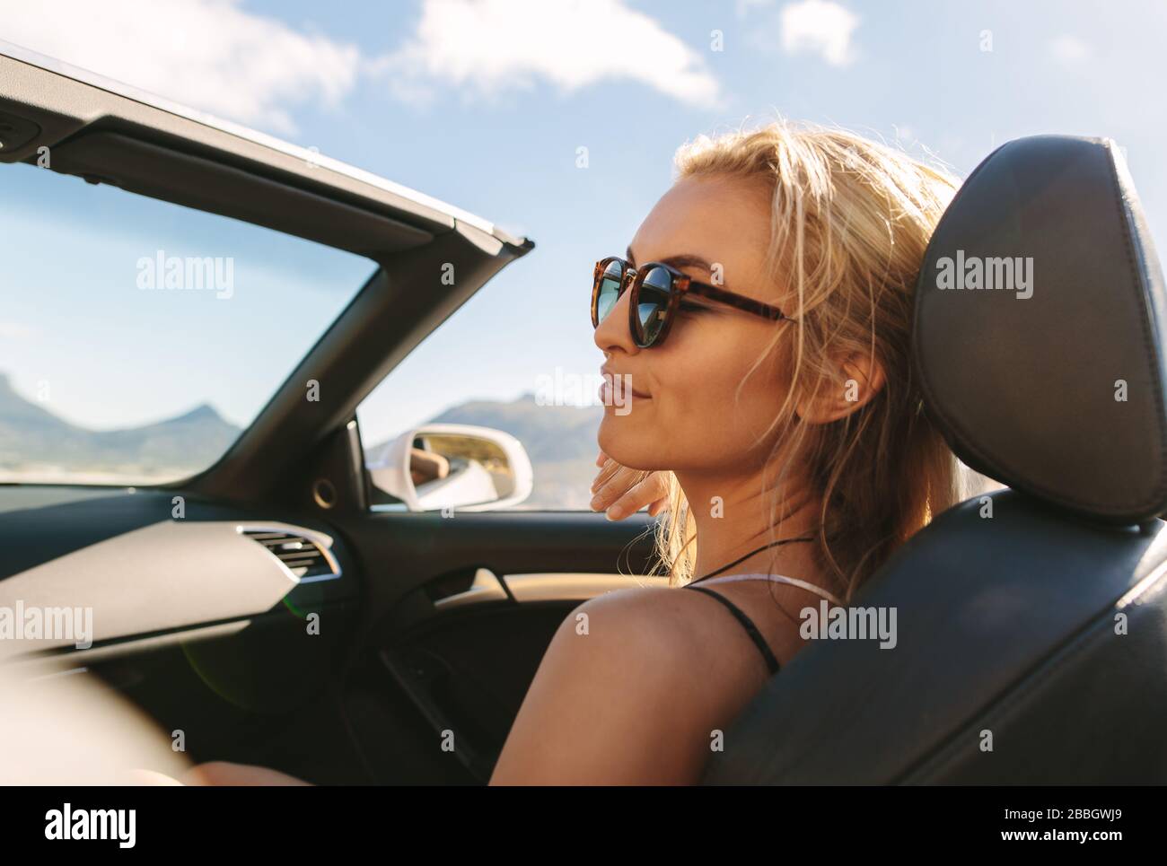 Woman on roadtrip travelling by a car. Caucasian female wearing sunglasses sitting relaxed in the passenger seat of a convertible car. Stock Photo