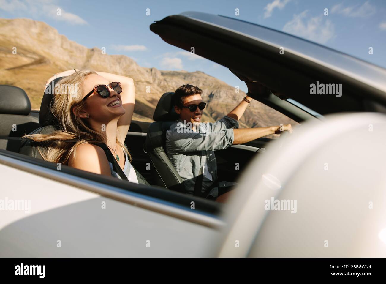 Cheerful couple having fun on a road trip. Young man driving convertible car with woman smiling on passenger seat. Stock Photo