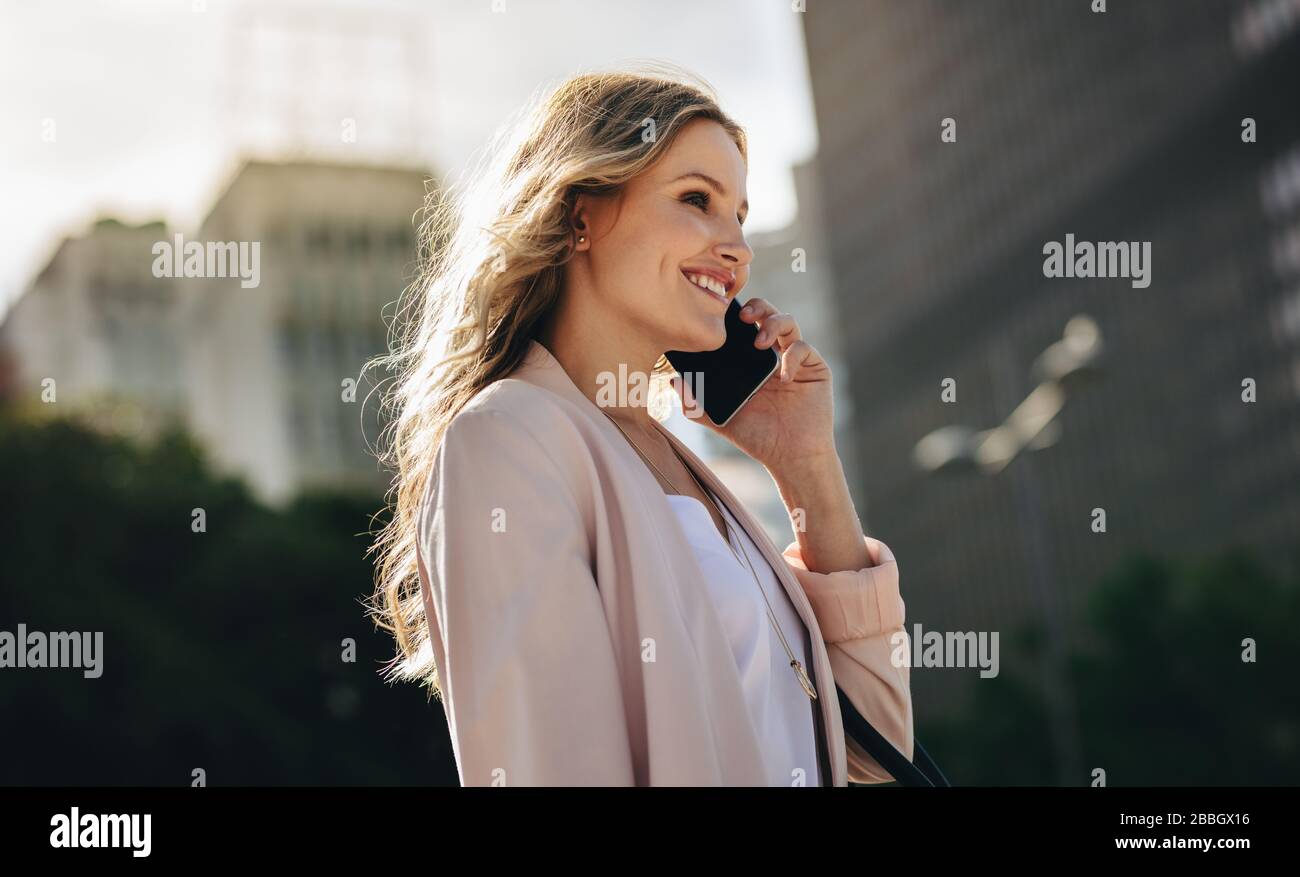 businesswoman using cell phone walking outside. Smiling female executive walking in the city making a phone call. Stock Photo