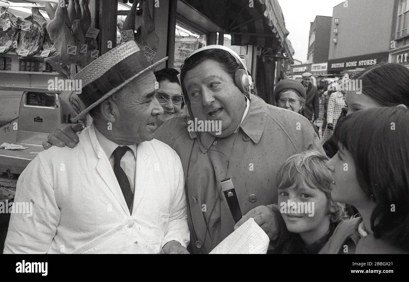 1970s, popular, man of the people, radio presenter and reporter, Monty Modlyn in a high street talking to a local shopkeeper, a butcher, surrounded by children in a South London market area, England, UK. Born into a Jewish family from Lambeth, Modlyn was known for his cockney accent and his broadcast forte was interviewing ordinary people, a technique that became known as 'Vox Pop'. Addressing people with the words 'Ullo darlin', his east end working class persona saw him have conversations about anything and everything during a long and distinguished broadcasting career. Stock Photo