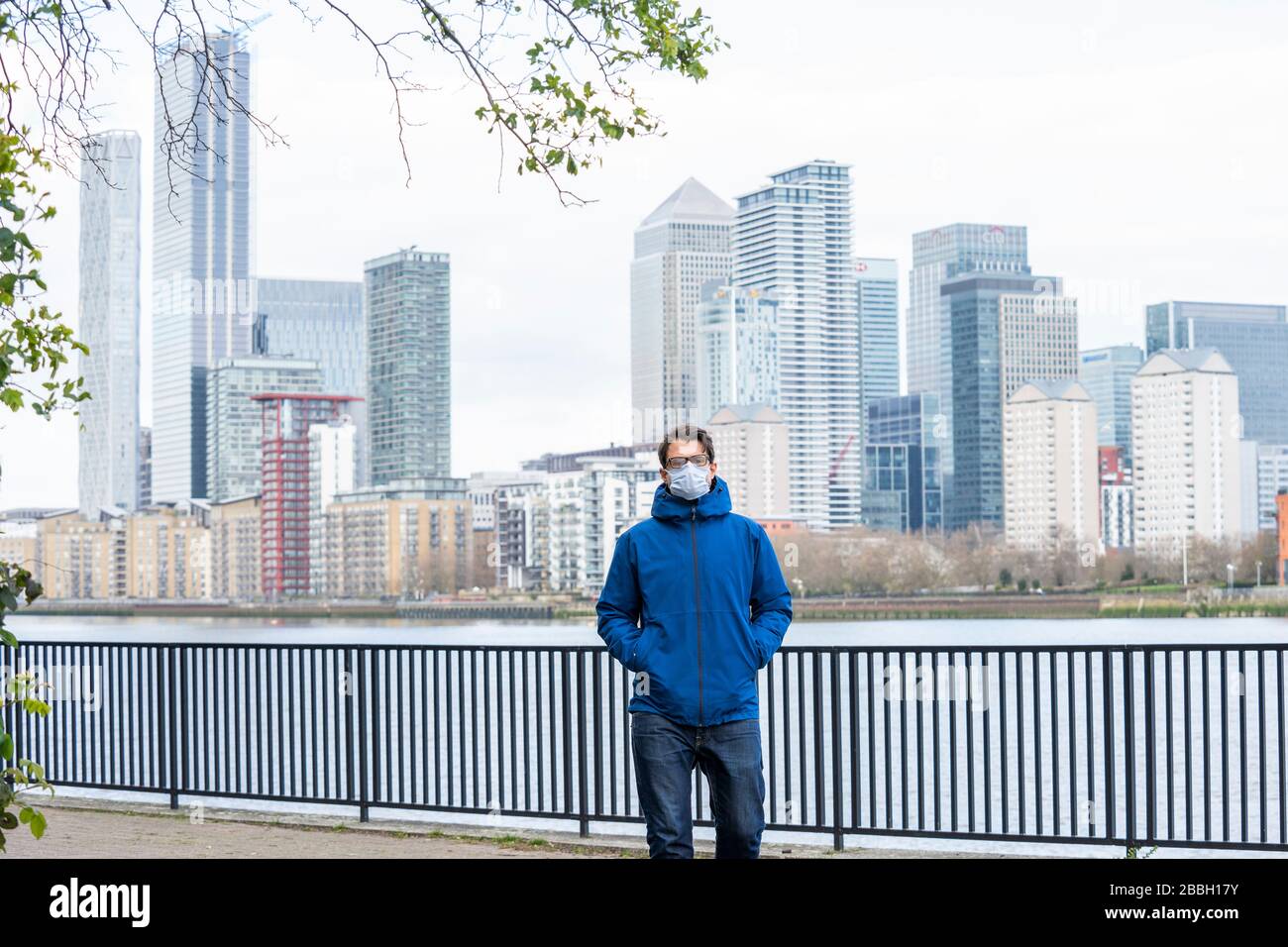 Mid 40's male wearing blue jacket and face mask walks along the River Thames, with Londons financial district and Canary Wharf in the background. Stock Photo