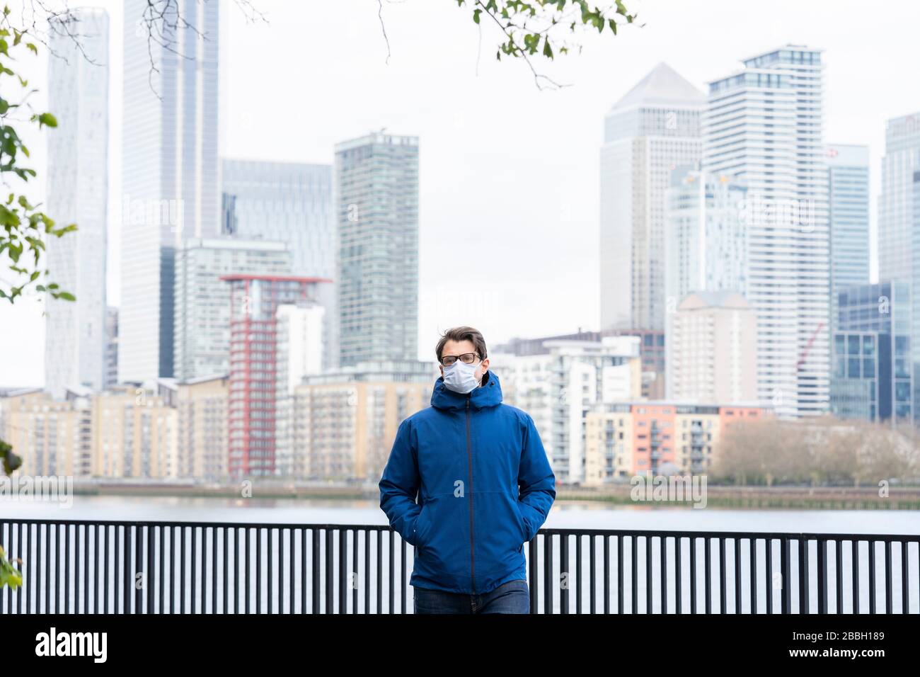 Mid 40's male wearing blue jacket and face mask walks along the River Thames, with Londons financial district and Canary Wharf in the background. Stock Photo