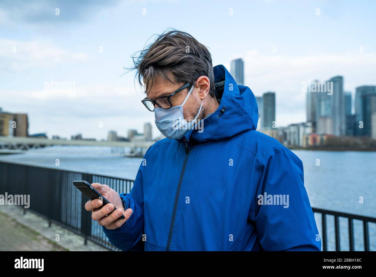 Mid 40's male in blue jacket, face mask and iPhone walks along the River Thames, with Londons financial district and Canary Wharf in the background. Stock Photo