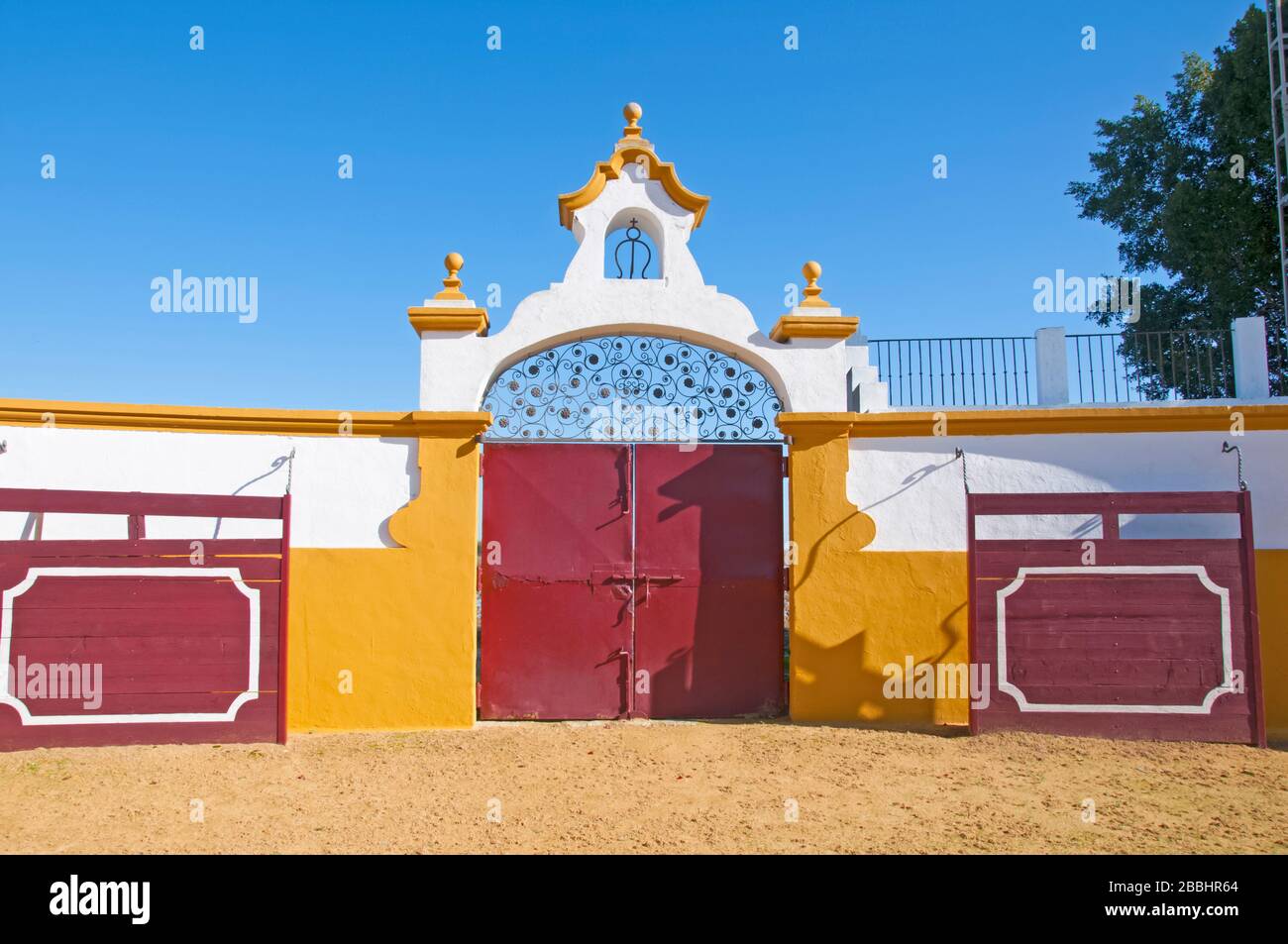 Big red gates in the bullring, white walls with orange decoration, two red barriers and sandy ground.  Isla Mayor, Spain Stock Photo