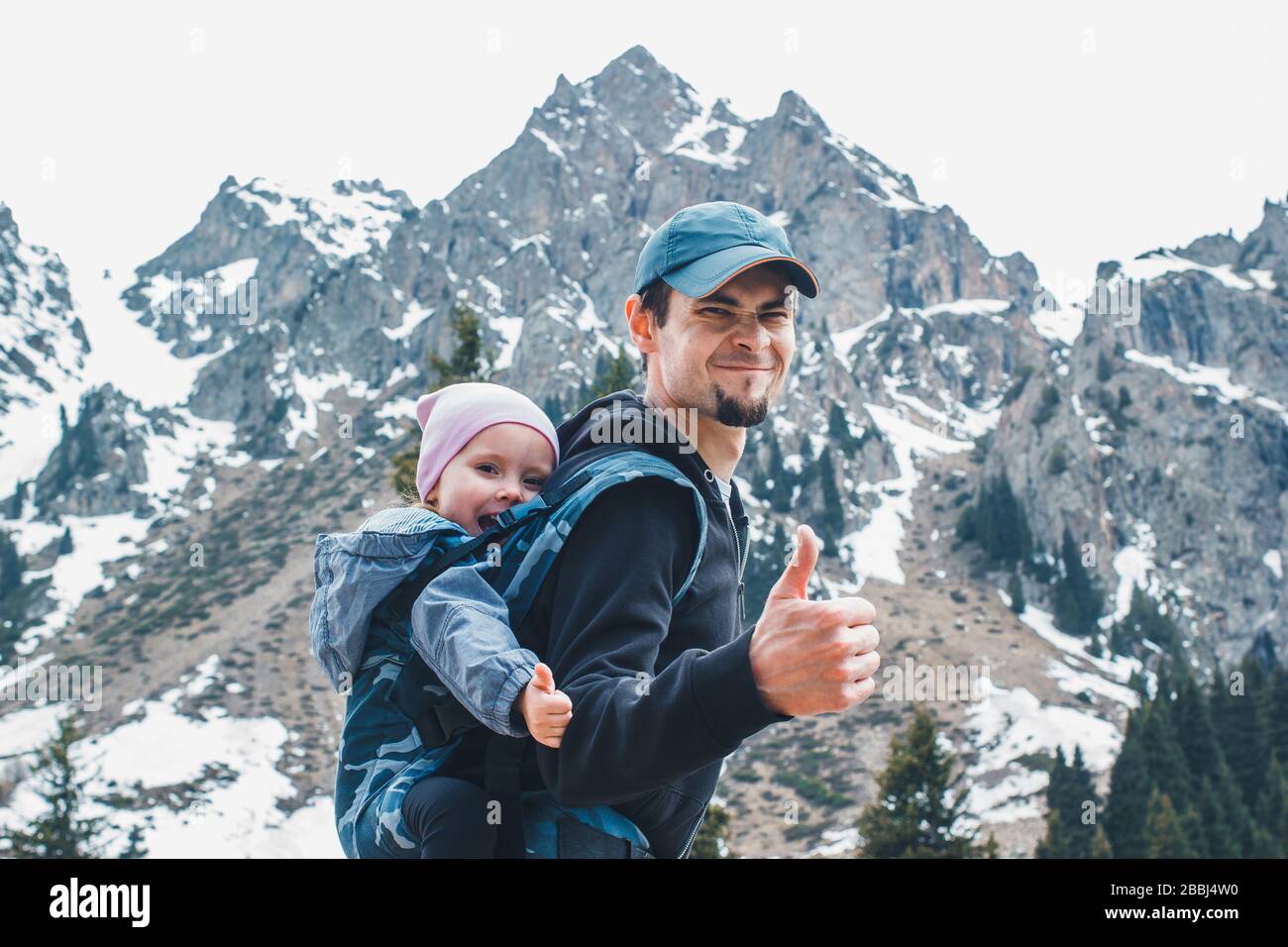 Attractive young father with her baby daughter in ergonomic baby carrier on back outside in mountain nature, show thumbs up. Stock Photo