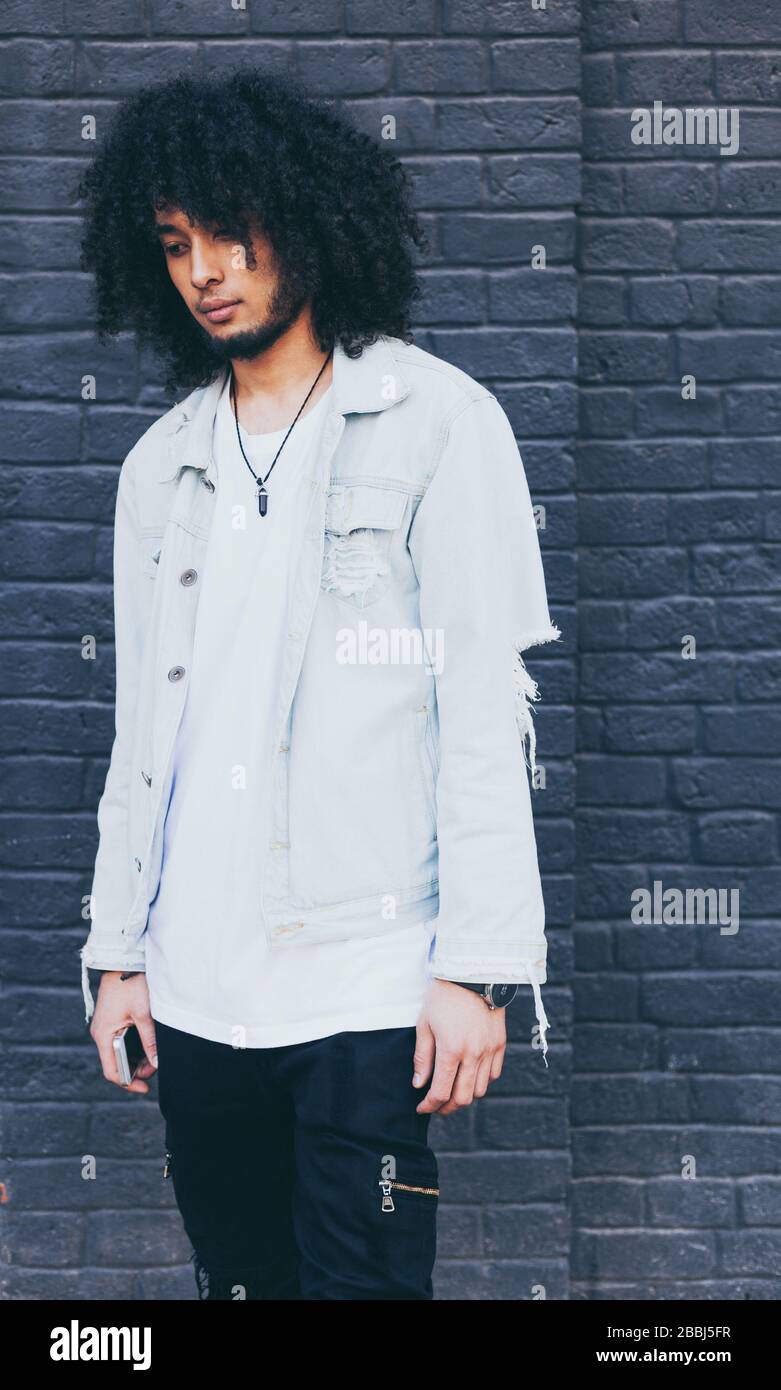Young black man with afro hair posing on graphite background. Outside. Dressed in a jeans jacket, black trousers. Fashion. Street style. Stock Photo
