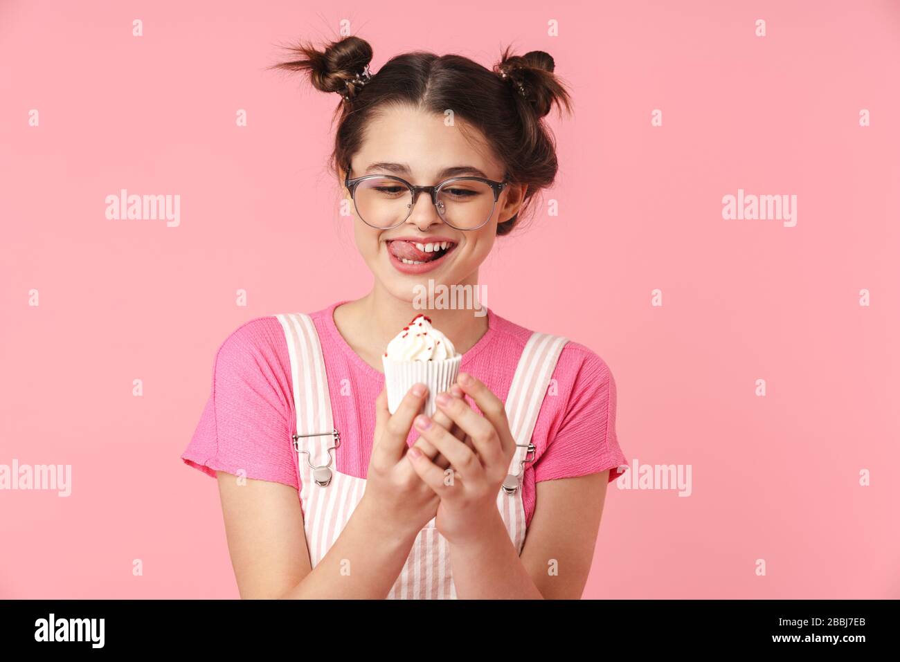 Photo of pleased charming girl in eyeglasses holding cake and licking her lips isolated over pink background Stock Photo