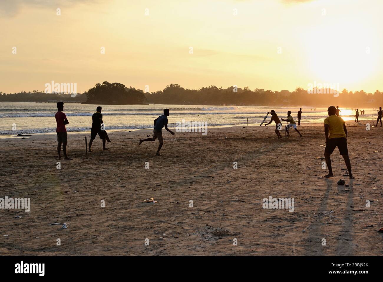 Young boys playing cricket game on a beach. Silhouettes of young  boys having fun on Sri Lanka beach. Golden sunset light with sunbeams in Weligama Stock Photo