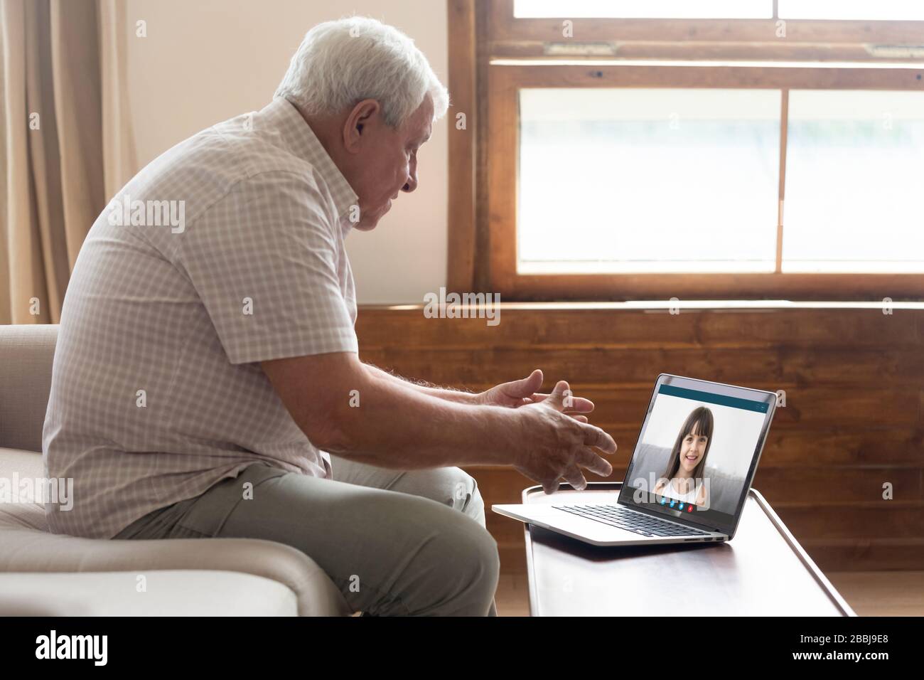 Senior grandfather sit on sofa quarantine at home speak talk on video call with smiling granddaughter, elderly grandparent use laptop go online commun Stock Photo