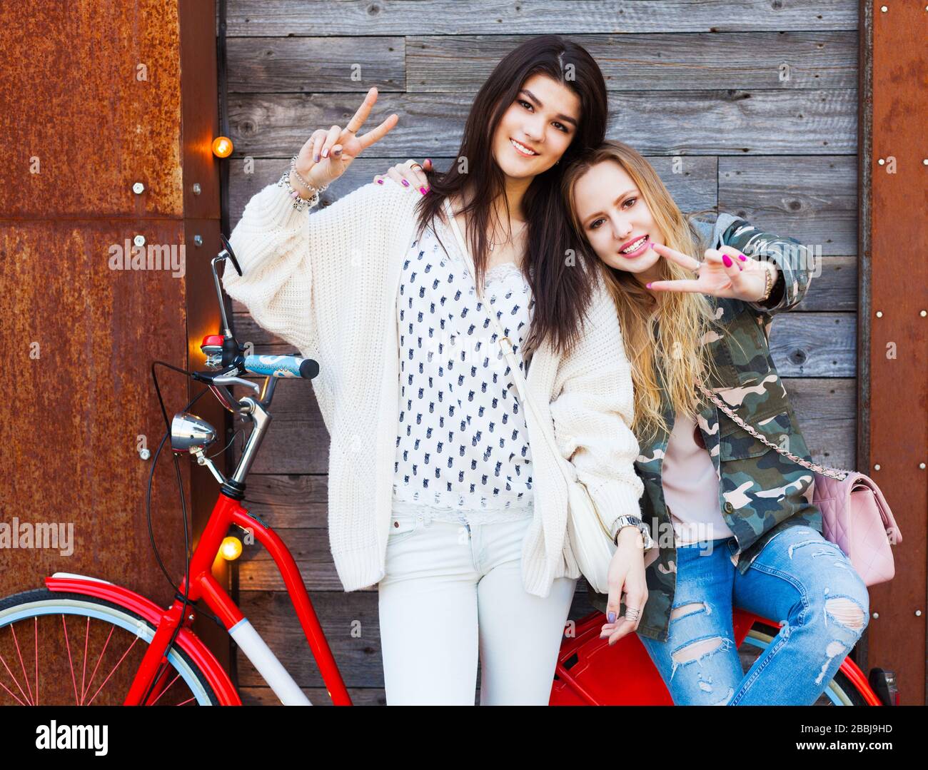 Two Trendy Fashionable Girls with red vintage Bike on old Wooden Background Show a sign of victory. Toned Photo. Modern Youth Lifestyle Concept. Stock Photo
