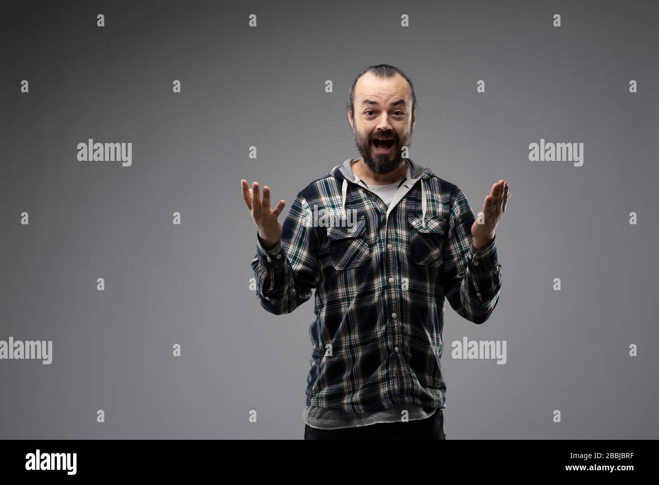 Happy bearded man in checked shirt showing surprised gesture with hands up in front of him, looking at camera. Half-length front portrait against grey Stock Photo