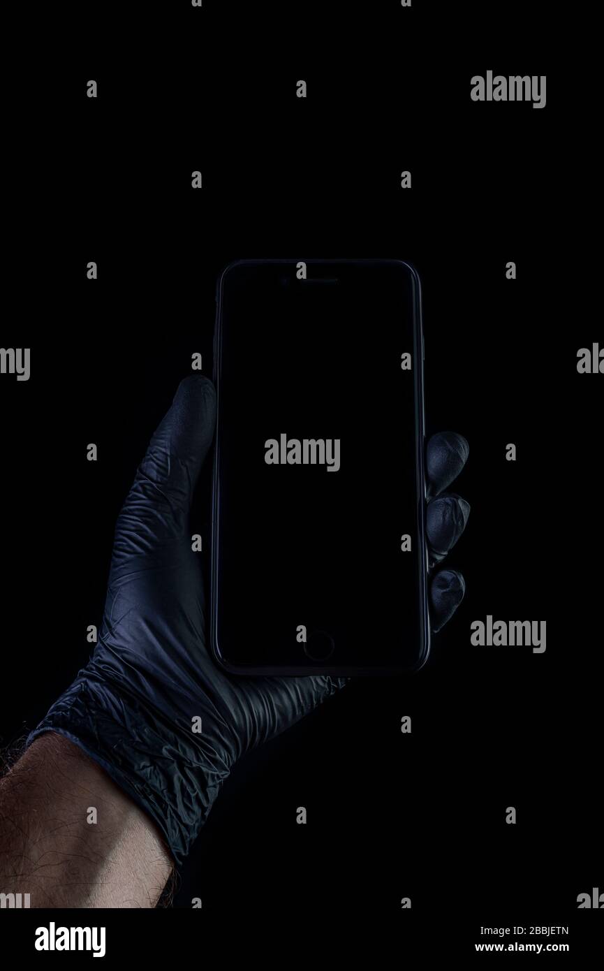 Hand holding a black phone isolated on a black background with a black gloves. Desinfection against coronavirus. Stock Photo