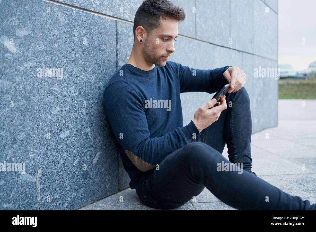 Side view of modern male in casual wear sitting near stone wall and br Stock Photo