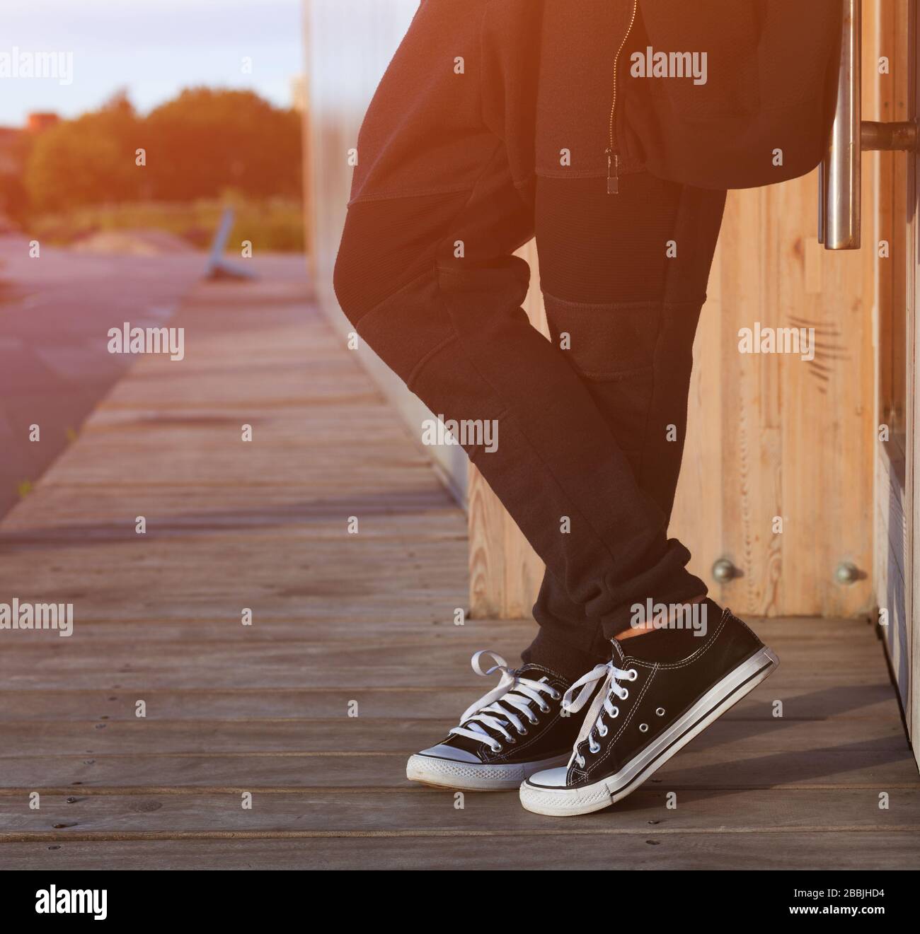A man in a fashionable black outfit posing at the sunset of a warm summer day in sneakers. Part of the body, close-up. Stock Photo