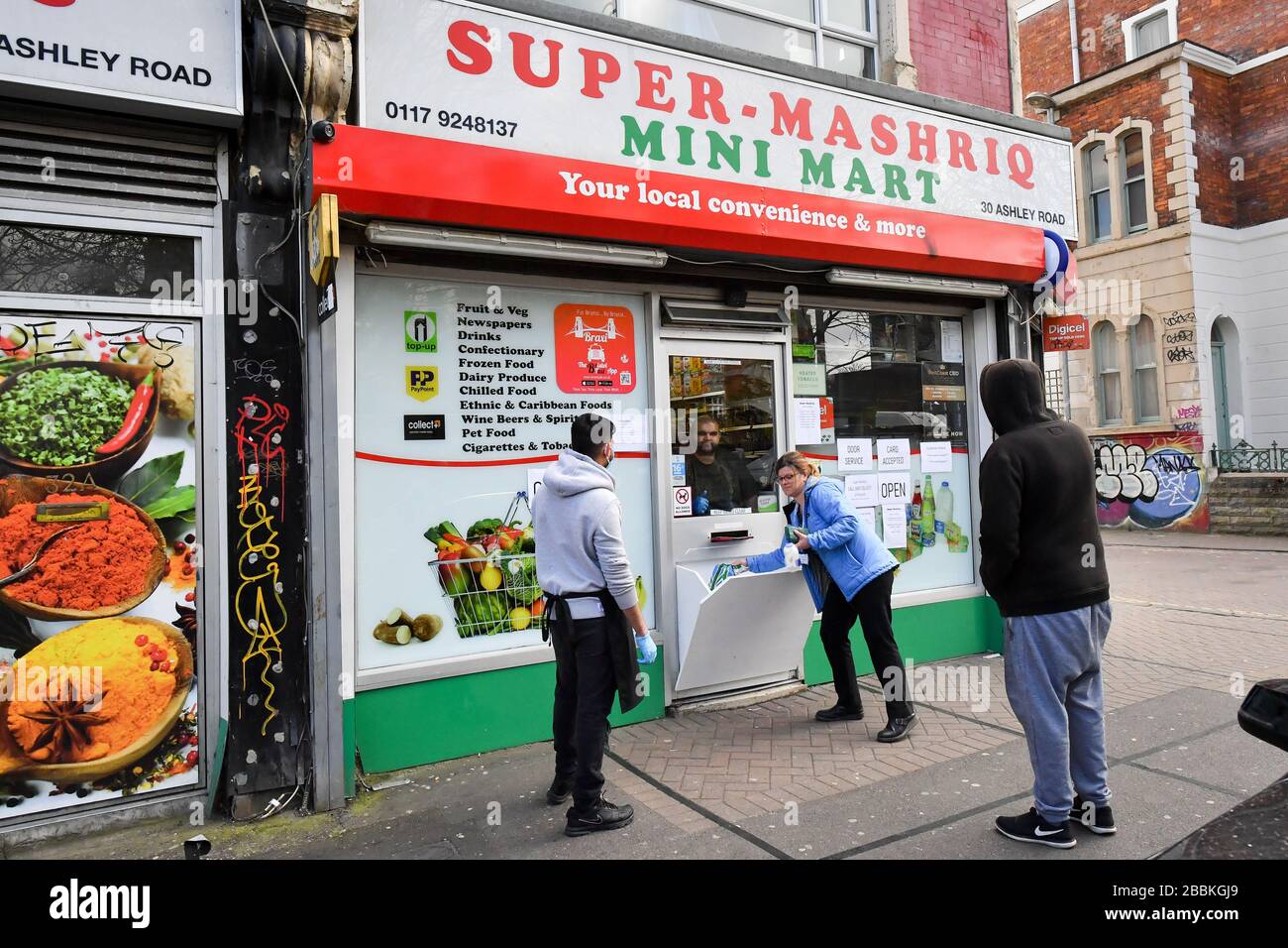 People buy food items from a modified shop door, that has been transformed into a serving hatch to avoid human contact, in St Pauls, Bristol, as the UK enters the second week of lockdown due to the global coronavirus pandemic. Stock Photo