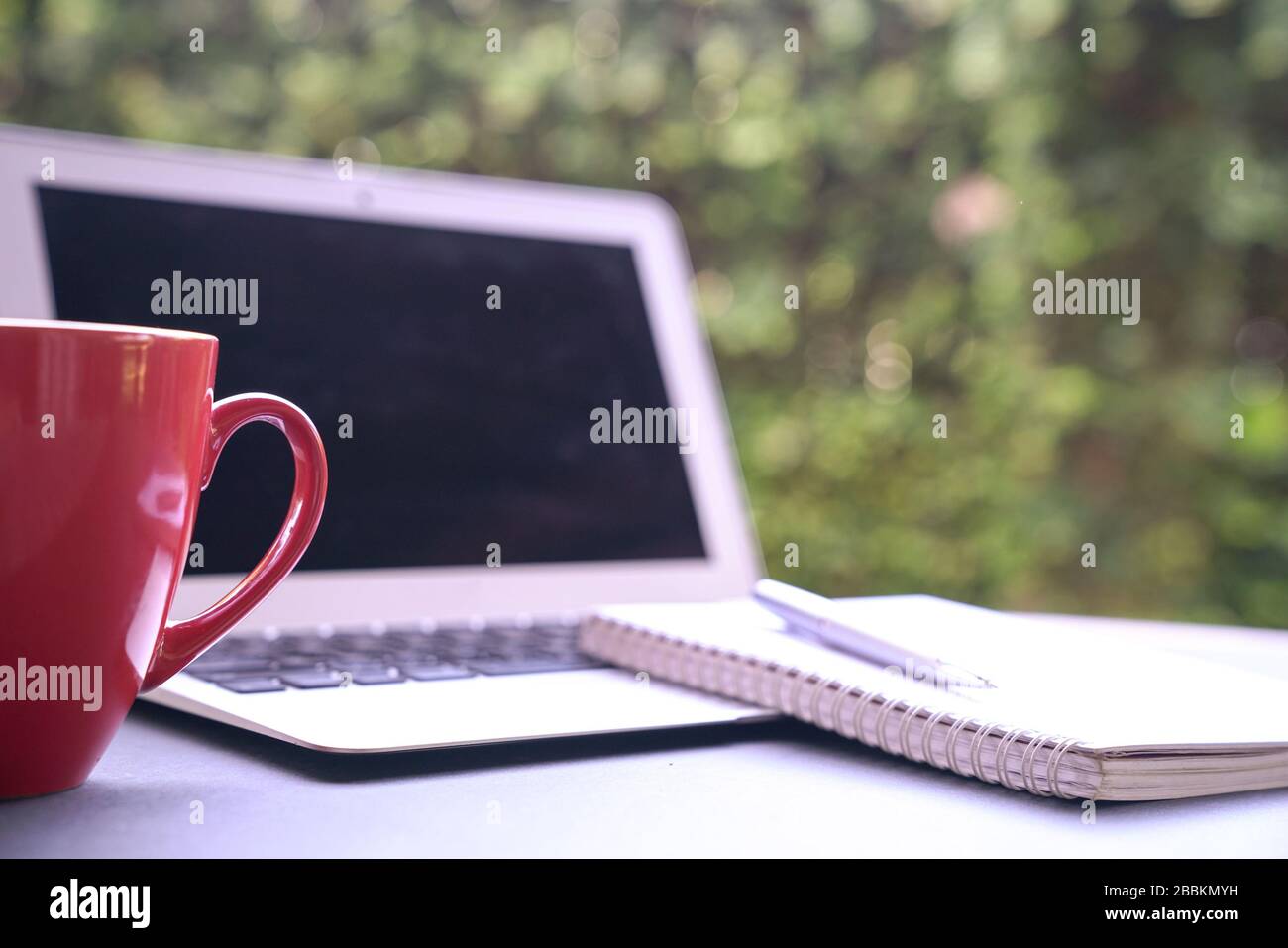 Computer laptop with notepad and coffee in red cup with green nature background. Work remotely or from home. Copy space. Stock Photo