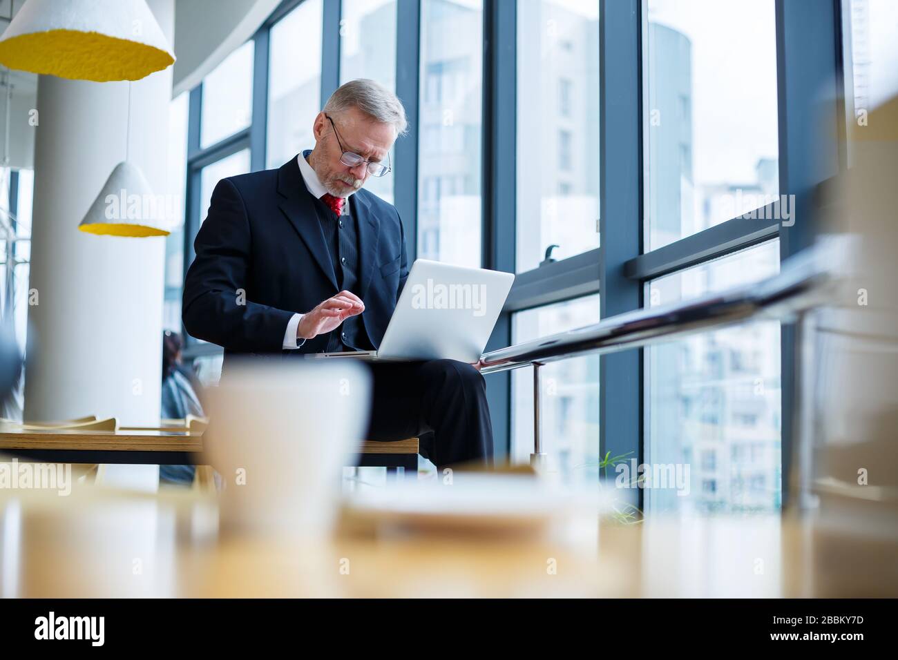 Smiling happy managing director thinks about his successful career development while standing with a laptop in his office near the background of a win Stock Photo