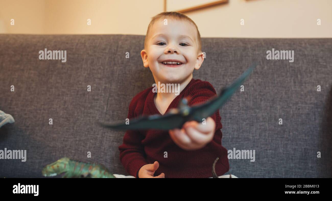 Caucasian small boy sitting on the sofa and playing with dinosaurs toys Stock Photo