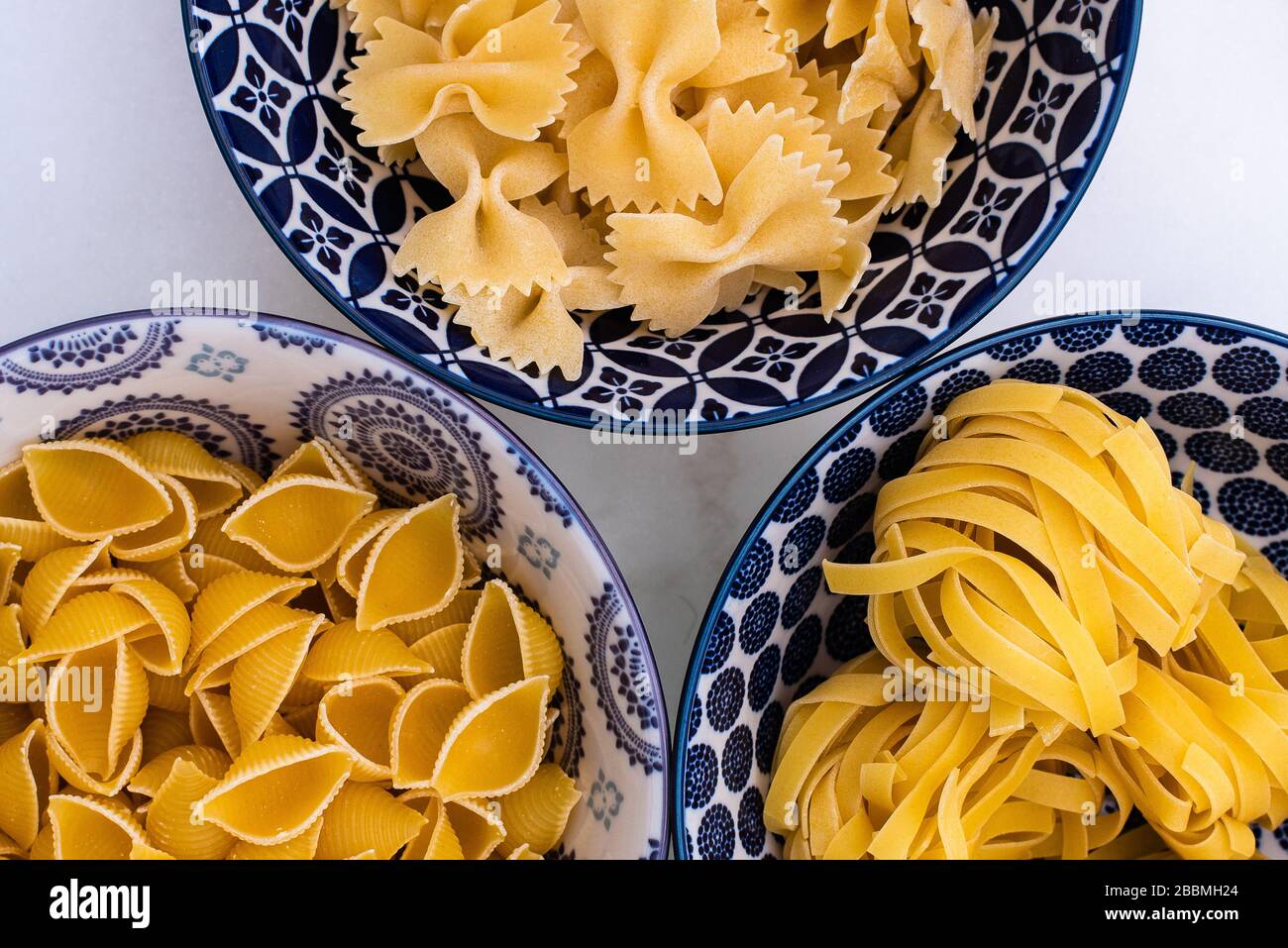 Different dry pasta shapes in patterned bowls on a white marble worktop