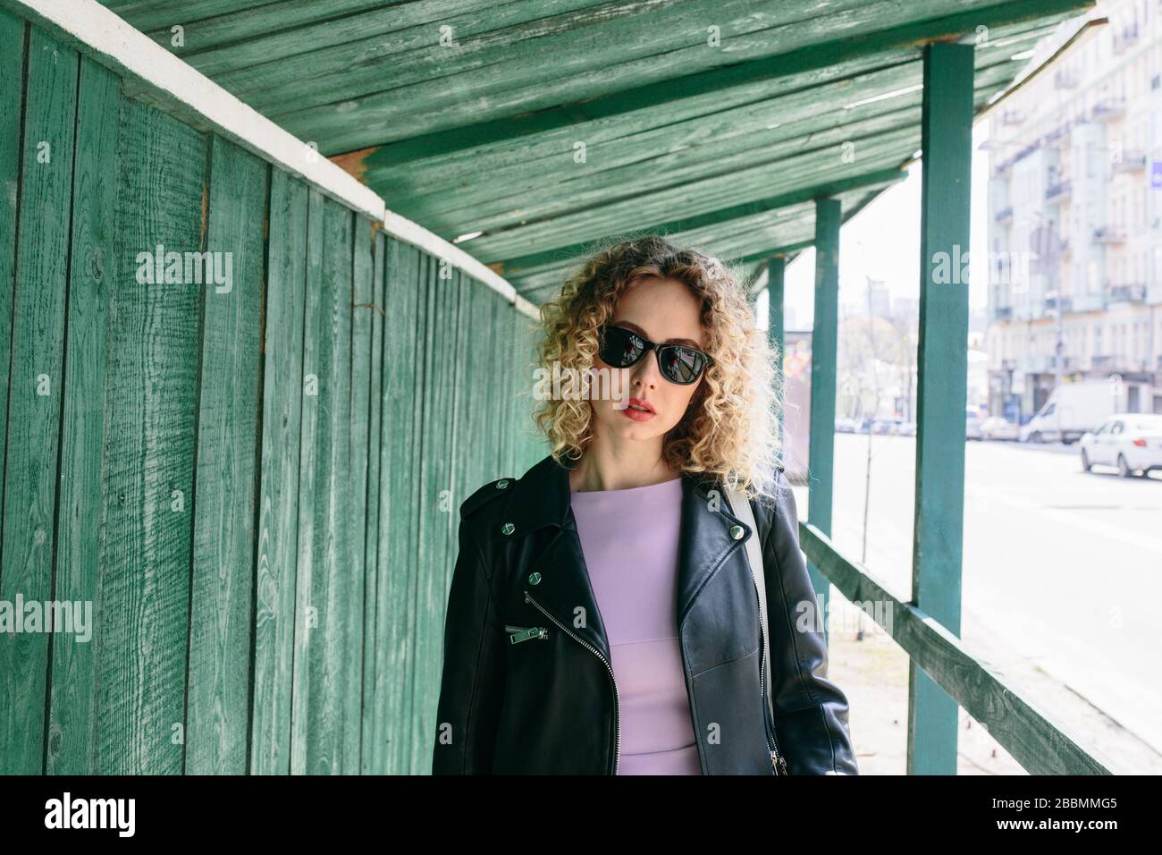 Portrait of beautiful women with curly long hair in black leather jacket and violet dress and black sun glasses in the green building transition Stock Photo