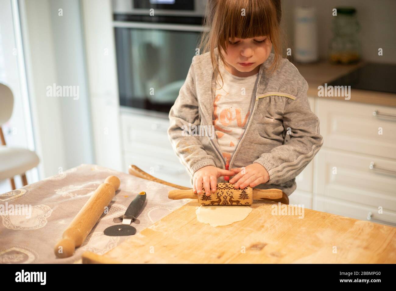 Child girl, 3 years old, in white kitchen, flattening pizza dough with a rolling pin on a wooden board. Lockdown activity idea. Stock Photo
